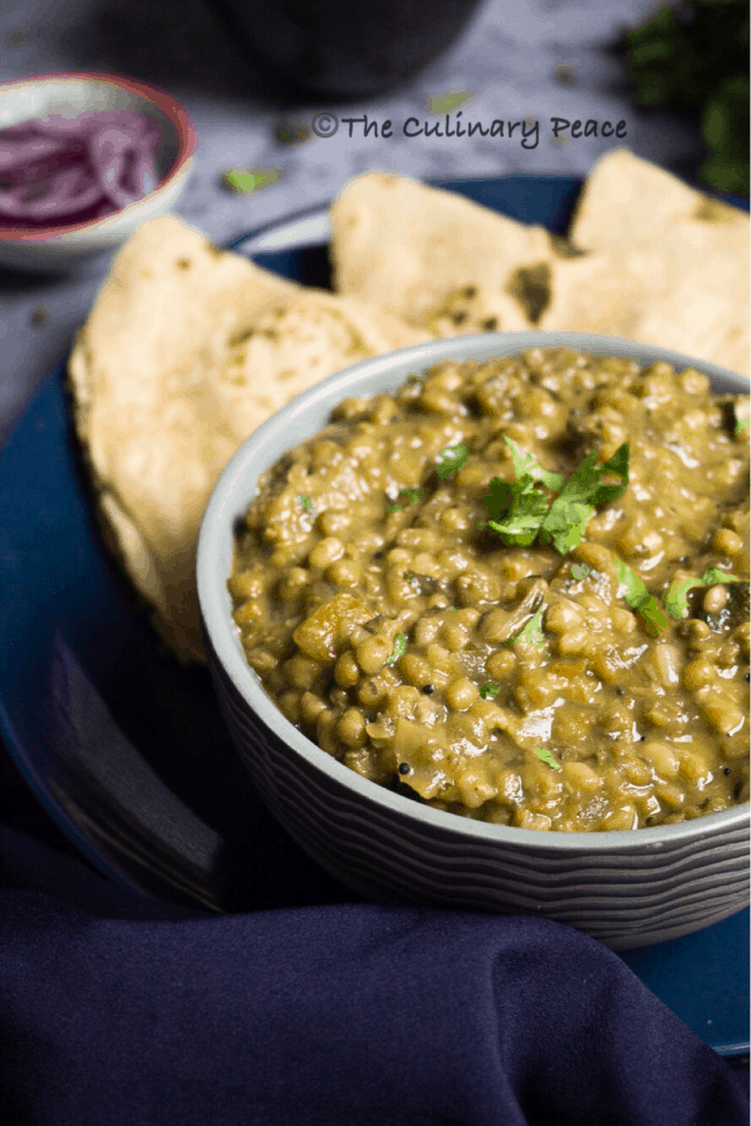 A healthy Indian curry recipe made of green gram with cilantro served inside a grey color bowl placed on a blue plate along with chapati against a grey background