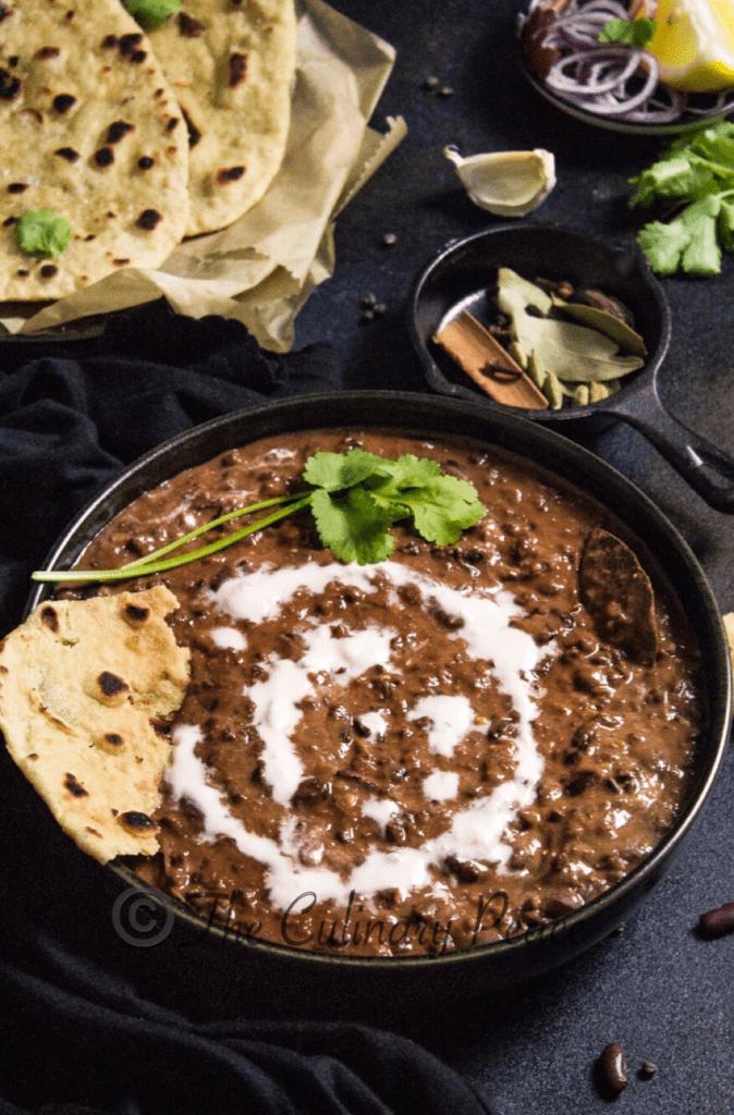 Creamy healthy Indian curry made of Kidney Beans and Black Lentils garnished with cilantro, cream served in a dish along with naan bread against a black background
