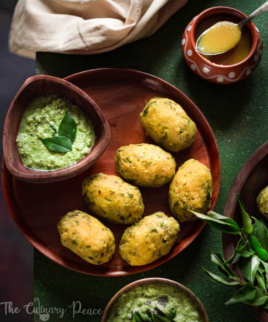 Flatlay of nuchinunde served on a brown wooden plate with a bowl of coriander coconut chutney and ghee on a green background