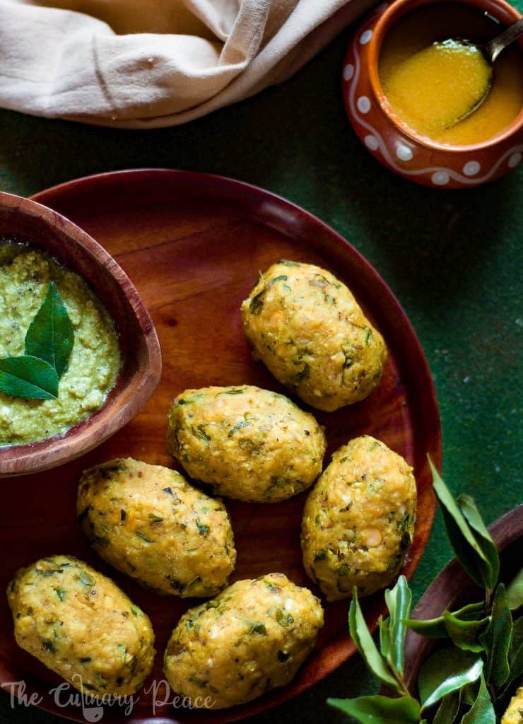 Flatlay of nuchinunde served on a brown wooden plate with a bowl of coriander coconut chutney and ghee on a green background