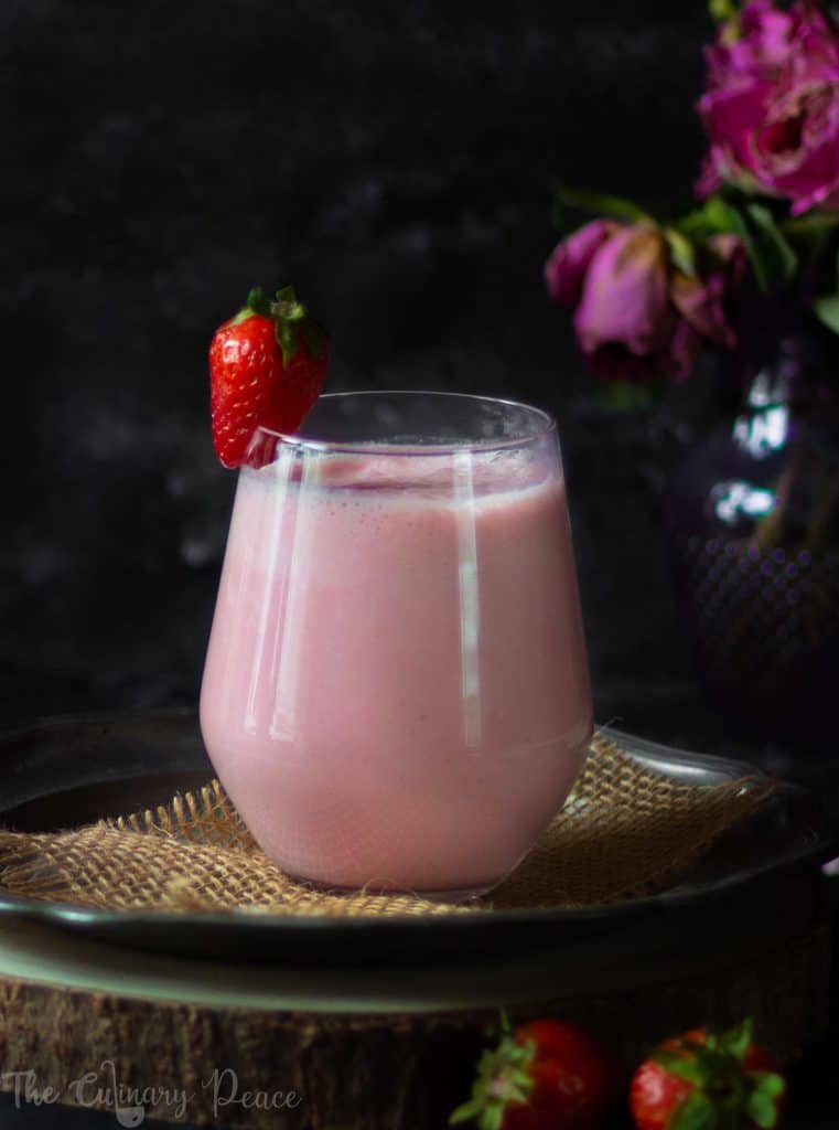 A picture of pink colored strawberry lassi served in a glass with fresh strawberries on a plate against a dark background.